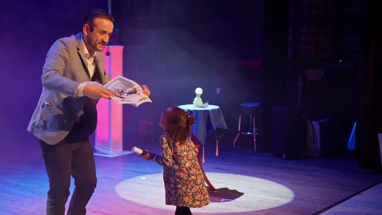 Image de l’extrait : sur scène, sous un rond de lumière, Tyson Dumas présente un tour à une petite spectatrice