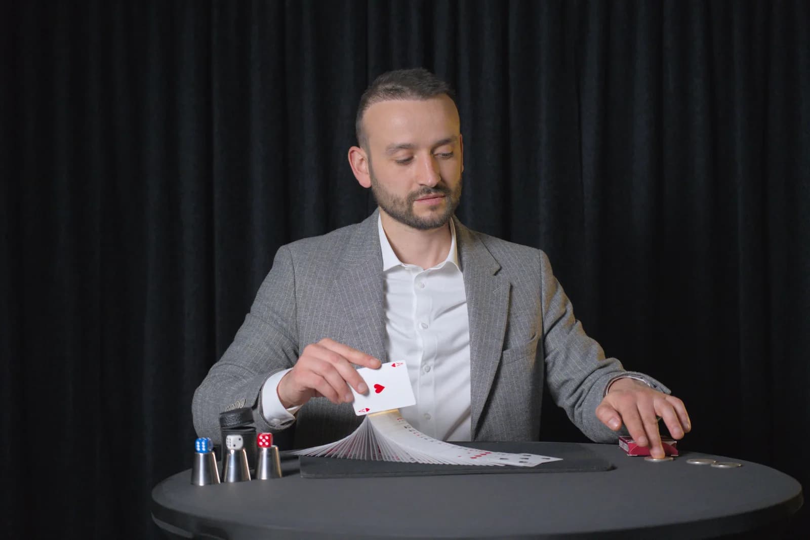 Tyson Dumas, magicien-mentaliste, assis derrière sa table de magie en costume gris à rayures fines, présentant l'As de cœur devant un jeu de cartes déployé en éventail